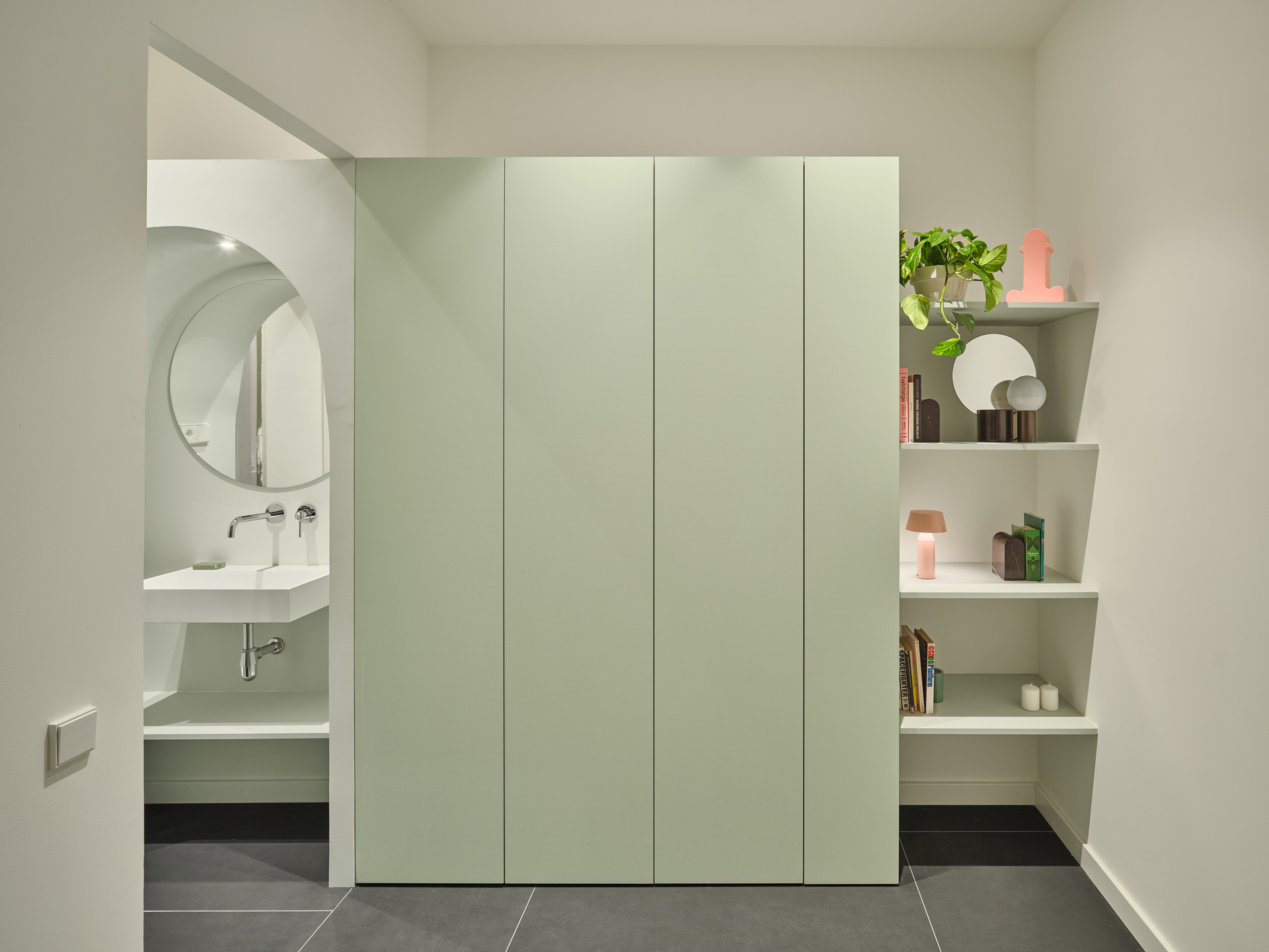 Bedroom circulation area in CRÜ’s “La Dolors” renovation, featuring a soft green built-in volume, open shelving, and a glimpse of the bathroom with a circular mirror and wall-mounted sink.
