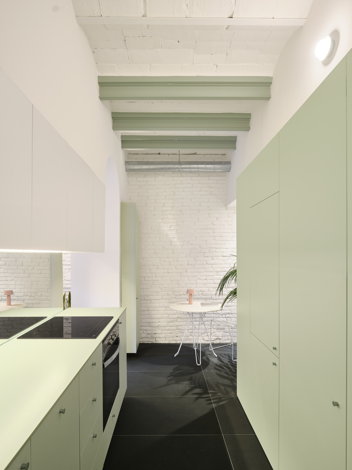 Kitchen and dining area in CRÜ’s “La Dolors” renovation, featuring pale green cabinetry, an island with induction cooktop, whitewashed brick walls, and exposed green beams.