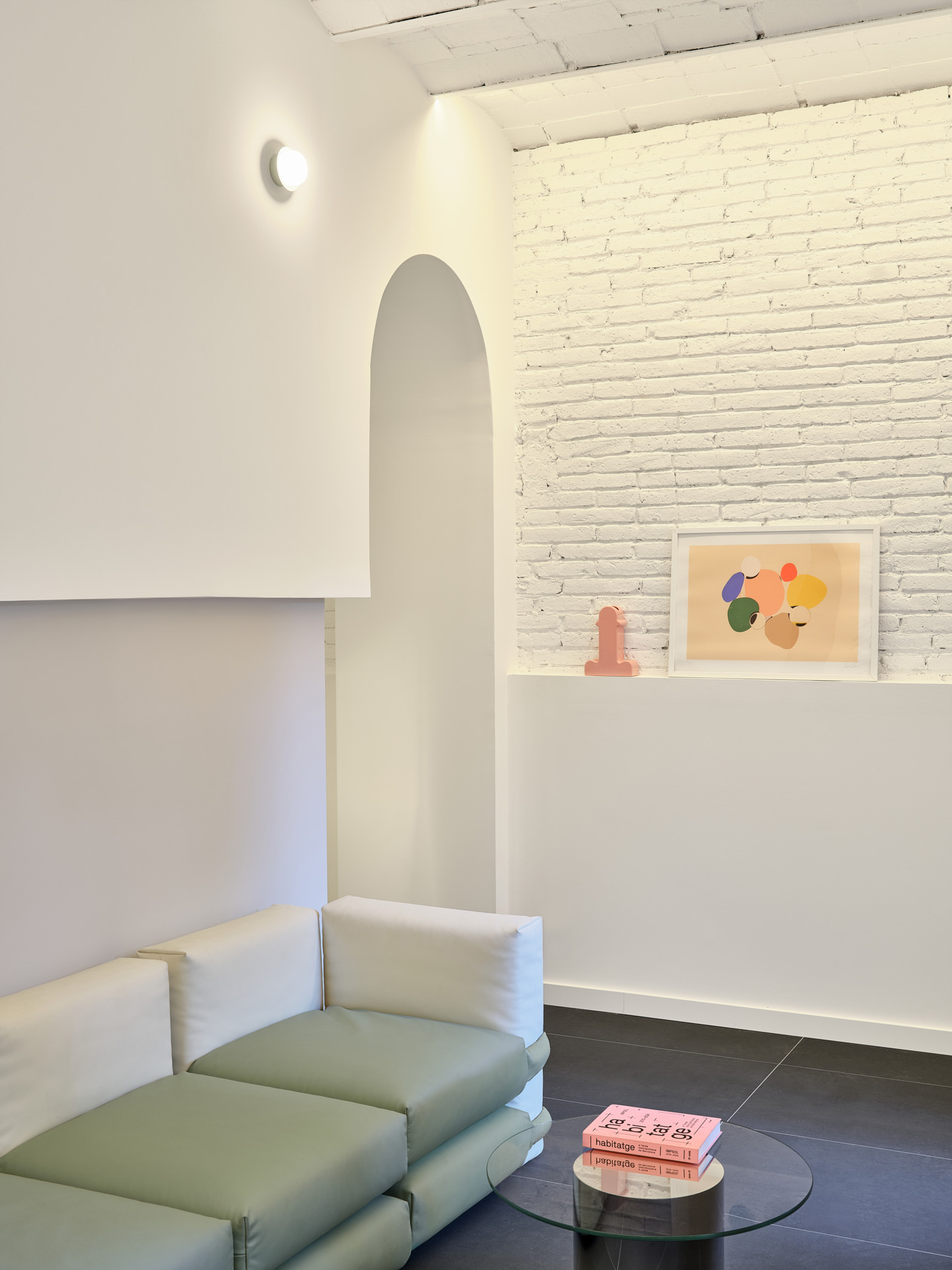: Living room of the “La Dolors” renovation by CRÜ, featuring a Muller Van Severen modular sofa in soft green tones, a whitewashed brick wall with artwork, and an arched opening revealing the restored ceramic structure.