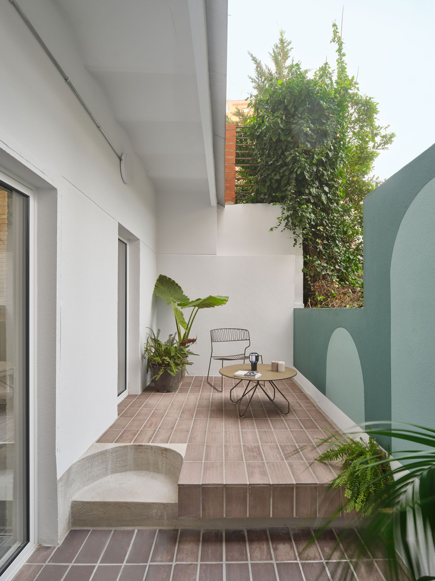 Terrace in CRÜ’s “La Dolors” renovation featuring a stepped ceramic platform, minimalist outdoor furniture, lush greenery, and a green wall with arched motifs